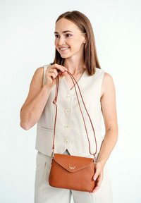Hnědá kožená crossbody taška s klopovým zavřením, zdobená logem a odnímatelným ramenním popruhem, kterou drží osoba ve světlé vestě.