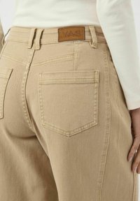 Béžové denimové kalhoty, které nosí osoba v bílém žebrovaném topu s dlouhým rukávem, ukazující zadní kapsu a hnědou koženou nášivku značky Y.A.S.