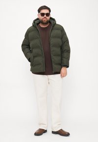 Man wearing dark green puffer jacket, brown shirt, white pants, brown boots, and sunglasses standing against plain white background.