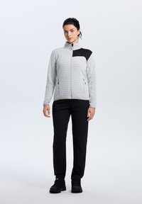Chaqueta de zip gris claro con un patrón texturizado y un acento negro, combinada con pantalones negros y calzado negro, de pie contra un fondo neutro.