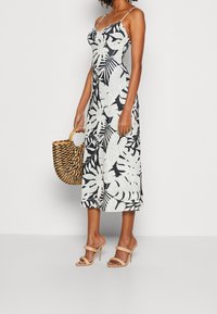 Robe maxi avec un motif de feuilles noir et blanc, bretelles spaghetti et un corsage ajusté ; associée à un sac en osier et des sandales à talons nude.