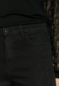 Jean en denim noir avec une texture lisse, une coupe slim, et des embellissements de petites clous sur la zone des poches avant, associés à un haut noir.