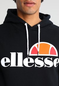 Svart hoodie med en stor vit "ellesse" logotyp och en orange och röd halvcirkel-grafik. Vita dragskommar med silvriga spetsar.