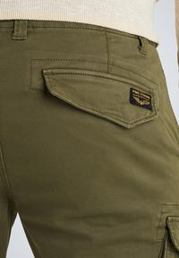 Olivno zeleni vojaški hlače z gladko teksturo, z bočno žepom in črnim etiketnim oznako z rumenim besedilom, nameščenim na zadnji strani.