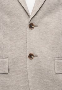 Μπεζ υφασμάτινο σακάκι με δύο καφέ κουμπιά και τσέπες με καπάκι, με γιακά λαιμού τύπου notch lapel.