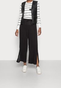 Zwarte wijde broek met zijsplitten, gecombineerd met een zwart leren gilet en een witte lange mouwen gestreepte blouse. Witte sneakers maken de look af.