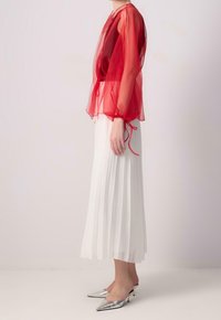 Rote transparente Bluse mit Puffärmeln und Bindebändern, kombiniert mit einem weißen plissierten Rock und silbernen spitzen Pumps. Strukturierte, leichte Materialien.