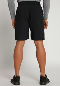 Shorts de sport noirs avec une taille élastique, arrivant au milieu de la cuisse, associés à des baskets noires sur un fond neutre.