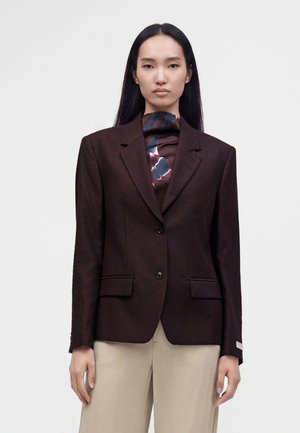 Femme aux longs cheveux noirs portant un blazer marron foncé, une écharpe à motif et un pantalon beige clair, debout devant un fond uni.