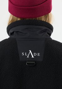 Giacca in pile nera con colletto alto, dotata di una toppa in tessuto nero e un logo bianco. Il modello indossa un cappello di lana bordeaux.