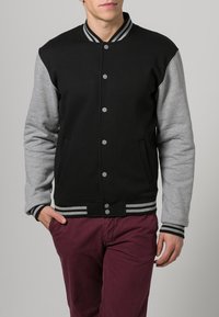Zwarte varsityjack met grijze mouwen, geribbelde kraag en gestreepte accenten. Voorzien van drukknoopsluiting en twee zijzakken.