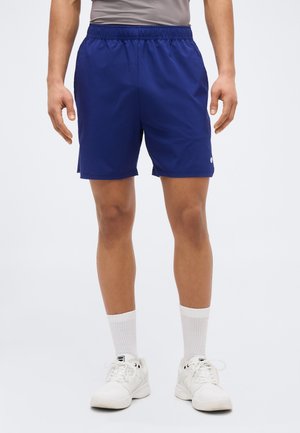 Hombre con pantalones cortos deportivos azules, calcetines blancos y zapatillas blancas, de pie con los brazos relajados a los lados contra un fondo liso.