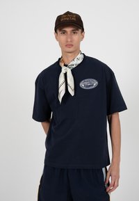 Jeune homme portant une casquette marron avec un texte, un t-shirt bleu marine avec un logo, un foulard à motifs et un short bleu marine assorti.