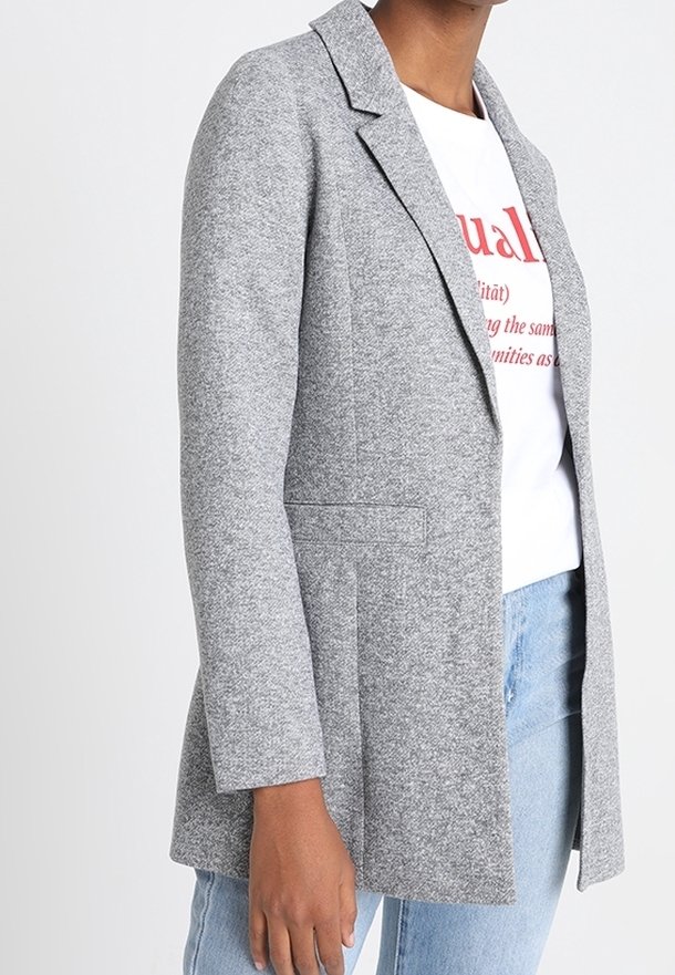 Grå kavaj med en texturerad yta, med ficklappar och två framfickor, bärs över en vit skjorta och blå jeans.