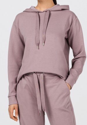Femme portant un sweat à capuche mauve et un pantalon de survêtement assorti avec cordons, main dans la poche, sur un fond clair uni.