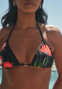 Bikini top negro con flores que presenta brillantes flores rojas y verdes, copas triangulares, tirantes finos y un cierre de metal plateado en la parte de atrás.