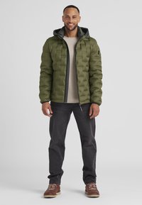 Grüne Pufferjacke mit Kapuze, mit gestepptem Design und einem Frontreißverschluss. Getragen über einem beigen Hemd mit dunklen Jeans und braunen Stiefeln.