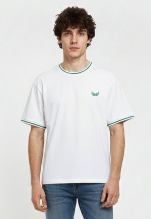 Hombre con cabello rizado que lleva una camiseta blanca de manga corta con ribete a rayas verdes y pequeño logo verde en el pecho, combinado con vaqueros azules.