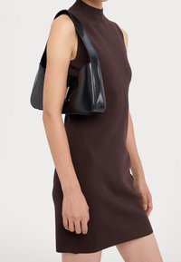 Svart, glänsande handväska med triangulär form och tjock axelrem, bärs över en mörkbrun, ärmlös, kroppsformad klänning.