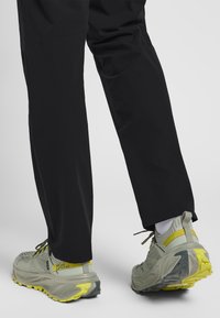 La chaussure présente une tige en maille respirante de couleur gris clair, avec une semelle épaisse en gris et jaune. Le pantalon est noir, avec une coupe ample et une texture subtile.