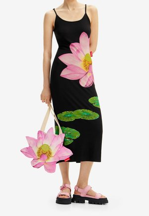 Femme portant une robe noire avec un grand motif de fleur de lotus rose et de feuilles vertes, tenant un sac assorti à la fleur de lotus et portant des sandales roses.