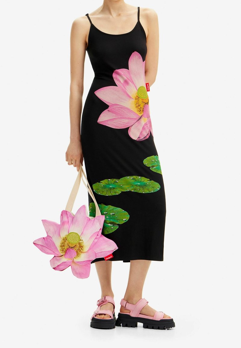 Femme portant une robe noire avec un grand motif de fleur de lotus rose et de feuilles vertes, tenant un sac assorti à la fleur de lotus et portant des sandales roses.