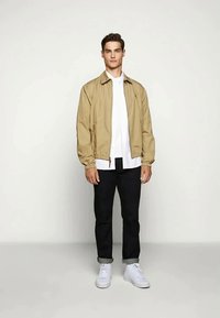 Young man standing, wearing beige zip jacket over white shirt, black rolled-up pants, and white sneakers against plain light background.