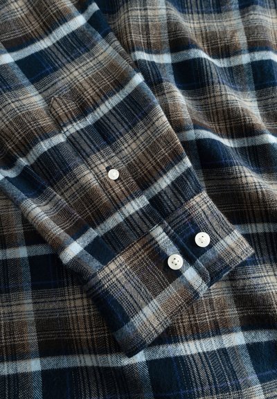 Chemise en flanelle avec un motif à carreaux bleu, marron et crème. Présente des poignets à boutons blancs et un tissu texturé. Apparence douce et chaude.