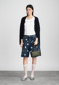 Bermuda shorts in denim con ricami floreali, abbinati a una maglietta bianca e a un cardigan blu navy, accessoriati con una borsa verde.