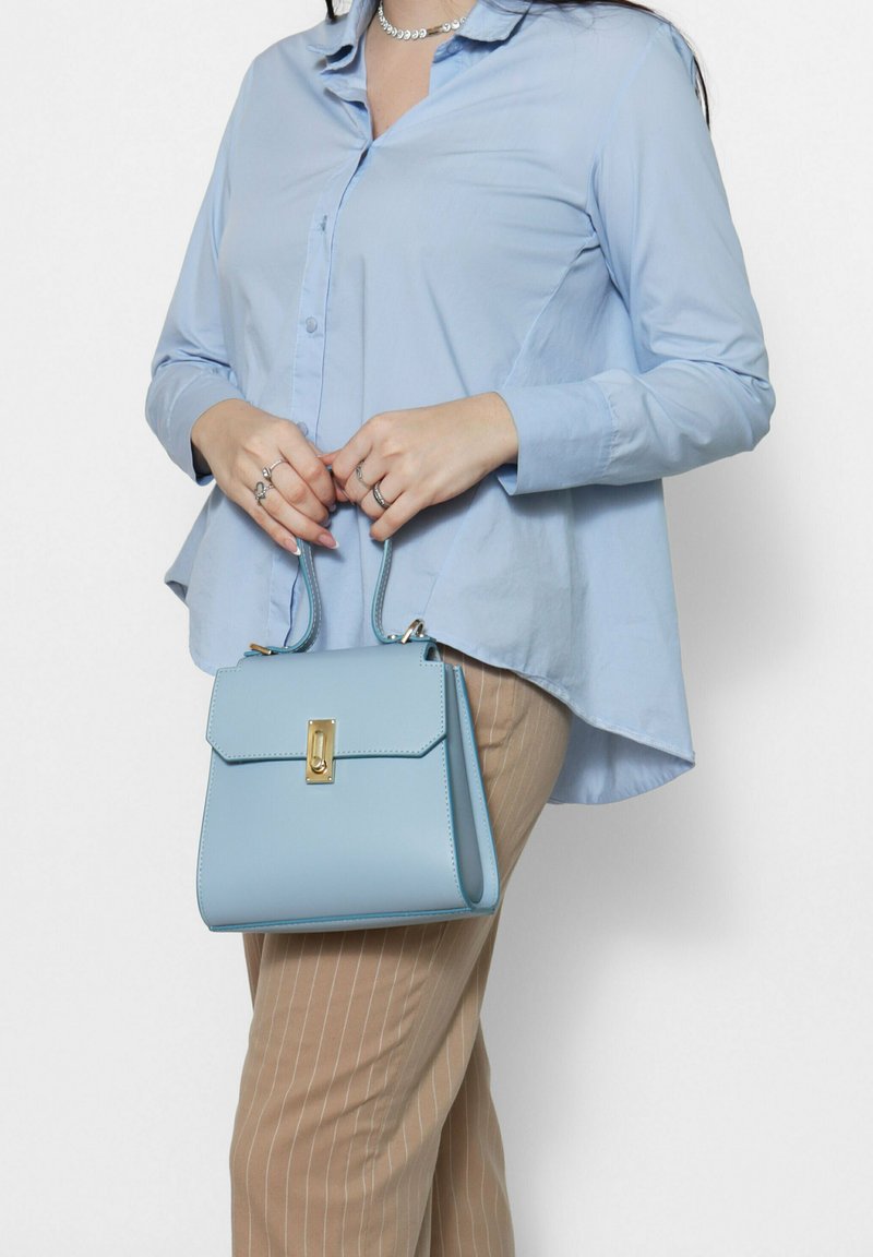 Lichtblauwe leren handtas met een gestructureerde vorm, gouden hardware, bovenhandvatten en een klep sluiting, aangevuld met een gestreepte beige broek en een blauwe shirt.