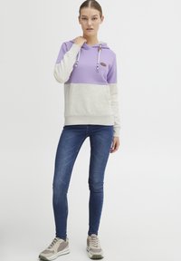 Felpa con cappuccio in viola e grigio chiaro, dal taglio rilassato, con lacci regolabili e una patch con logo. Abbinata a jeans skinny blu e sneakers.