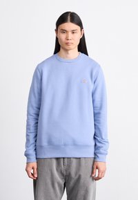 Light blue sweatshirt made of soft material, featuring a crew neck, ribbed cuffs, and a small logo patch on the chest. Worn with gray corduroy pants.