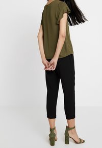 Olivgrünes, kurzärmeliges Blusenshirt mit einem Reißverschluss auf der Rückseite, kombiniert mit schwarzen, taillierten Hosen und grünen Riemchen-Pumps, die eine moderne Silhouette präsentieren.