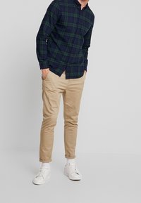 Camicia a maniche lunghe a quadri blu navy e verde, abbinata a pantaloni beige. Sneakers bianche con lacci. Fit casual, tessuto morbido, dettagli minimi.