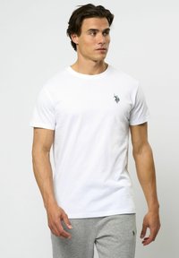 Vit bomulls t-shirt med korta ärmar och rundad halsringning, med en liten svart logotyp på vänstra bröstområdet. Slät textur.