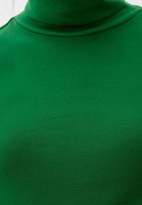 Grüner Rollkragenpullover mit gerippter Textur, figurbetontem Design und weichem Material. Nahaufnahme des Ausschnitts und des Oberkörperbereichs.