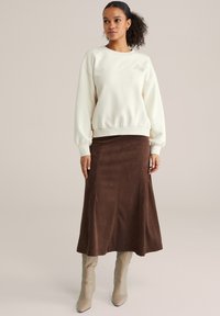 Frau mit lockigem Haar, die einen cremefarbenen Pullover, einen braunen Wildleder-Midirock und beige kniehohe Stiefel trägt, vor einem neutralen Hintergrund steht.