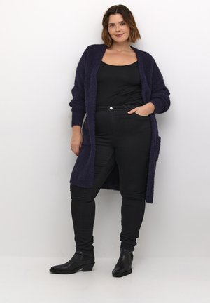 Lila, bolyhos kardigan zsebekkel, fekete felső és skinny farmer felett viselve. Hosszú ujjaival és laza szabásával rendelkezik. Fekete bokacsizmák.