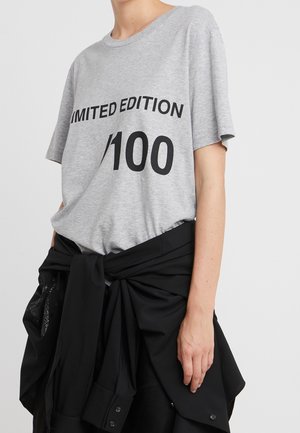 Osoba nosząca szarą koszulkę z napisem "LIMITED EDITION 1/100" i czarną kurtkę zawiązaną wokół talii na jednolitym tle.