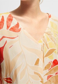 Kortærmet bluse med V-udskæring, der har et blomsterprint i bløde pastelfarver med fremtrædende blade i nuancer af rød, gul og fersken.