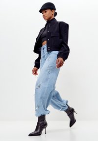 Zwarte cropped jas, getailleerde zwarte turtleneck, versleten lichtblauwe baggy jeans en zwarte spitse stiletto laarzen. Eenvoudige achtergrond.