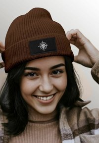 Brown ribbed beanie with a black patch featuring a compass design. The beanie is snug-fitting and textured, suitable for cold weather.