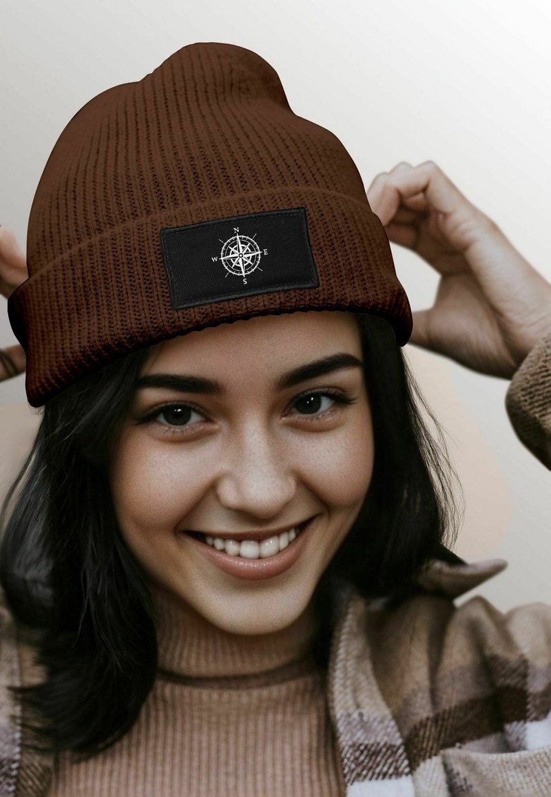 Brown ribbed beanie with a black patch featuring a compass design. The beanie is snug-fitting and textured, suitable for cold weather.