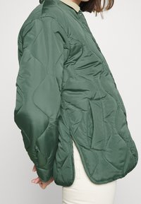 Giacca verde trapuntata con colletto rotondo, tasche laterali e un motivo tessile. Materiale leggero con una vestibilità leggermente ampia.