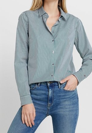 Kvinde iført en langærmet, hvid og mørkstribet skjorte med knapper, stoppet ned i blå jeans, med den ene hånd i lommen, stående mod en ensfarvet baggrund.