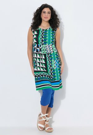 Frau mit lockigem dunklem Haar, die ein ärmelloses, mehrfarbiges geometrisches Kleid, blaue Leggings und weiße Keilsandaletten trägt und vor weißem Hintergrund steht.