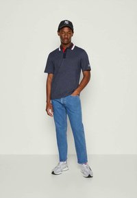 Homem com uma camisa polo azul-marinho, jeans claros, ténis cinzentos e um boné de beisebol preto, de pé contra um fundo claro e simples.