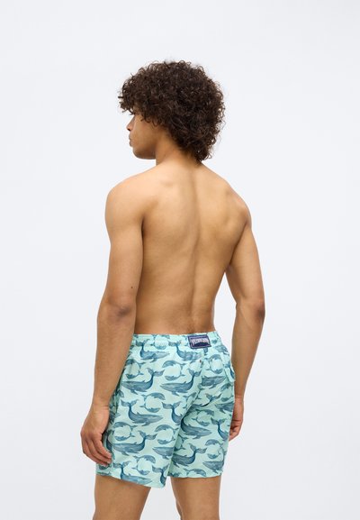 Muž bez trička s kučeravými vlasmi, otočený chrbtom, oblečený v svetlomodrých plavkách s tmavomodrými veľrybami na bielom pozadí.