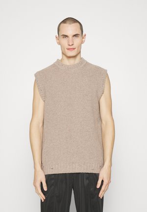 Πλεκτό αμάνικο πουλόβερ σε ανοιχτό μπεζ, με ψηλό λαιμό και ακατέργαστες λεπτομέρειες. Χωρίς μοτίβα, συνδυάζεται με σκούρες παντελόνες.