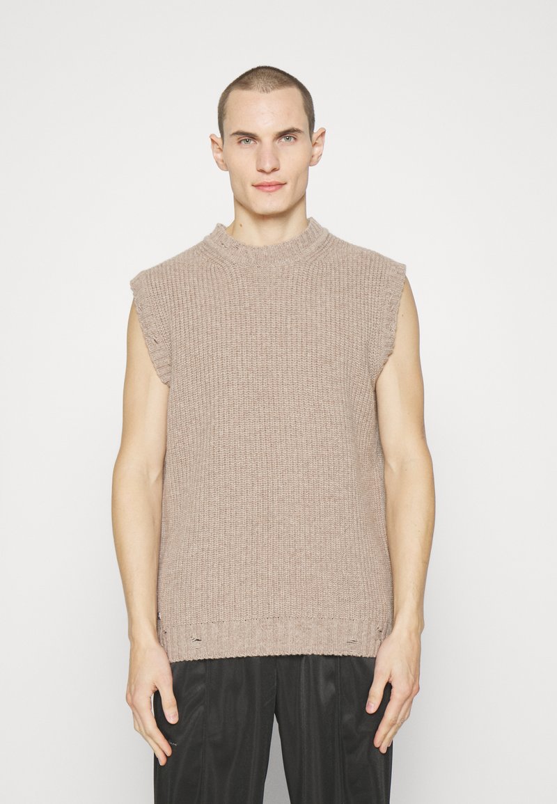 Πλεκτό αμάνικο πουλόβερ σε ανοιχτό μπεζ, με ψηλό λαιμό και ακατέργαστες λεπτομέρειες. Χωρίς μοτίβα, συνδυάζεται με σκούρες παντελόνες.